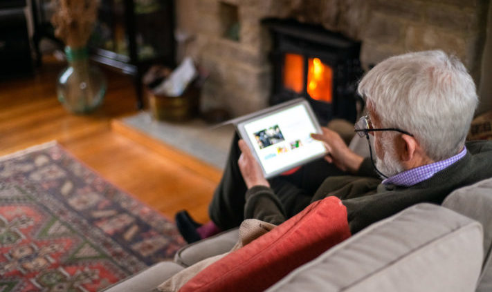 Older man relaxes on sofa by a fire, and looks pictures on his tablet.