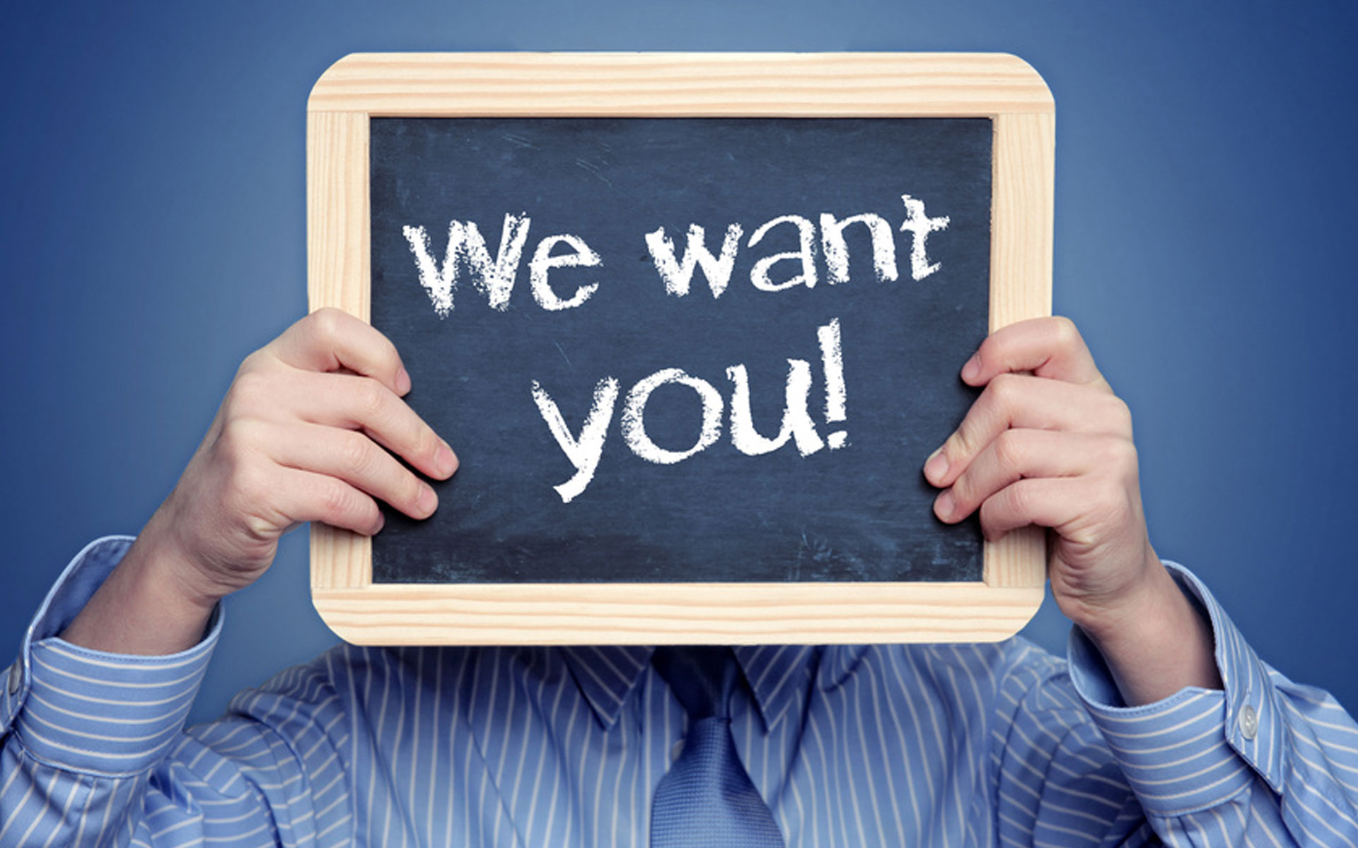 Man wearing blue shirt and tie holding a small chalkboard in front of his face that states: "We want you!"